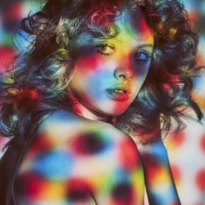 Naked by Guy Aroch, portrait of nude model in sixties style makeup and curly hair, covered in colorful light dots