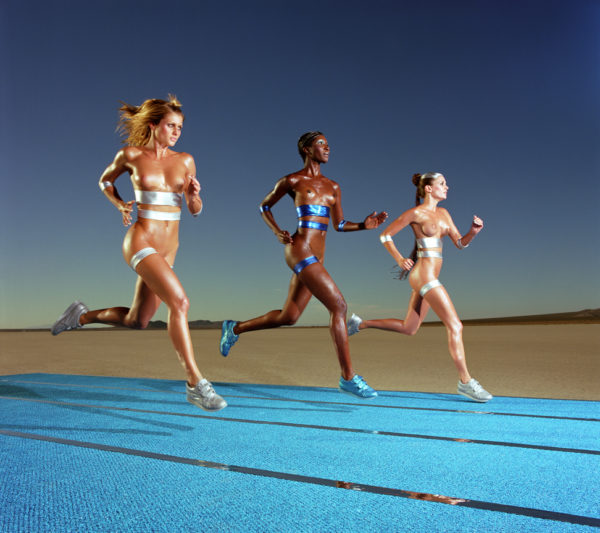 OLYMPIC RUNNING by Guido Argentini, three models in metallic sneakers and fabric stripes running on a blue carpet in the desert