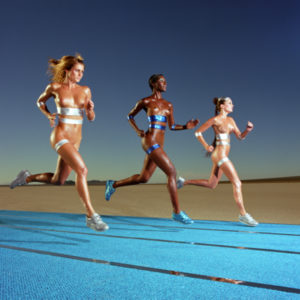 OLYMPIC RUNNING by Guido Argentini, three models in metallic sneakers and fabric stripes running on a blue carpet in the desert