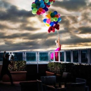 Away by David Drebin, model in pink dress standing on a balkony, holding colorful airballoons, a photographer behind her