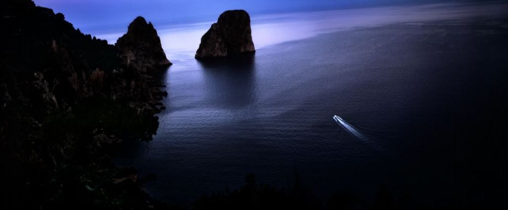 Capri Dreams by David Drebin, a boat on a quiet ocean in the dark next to rocky cliffs