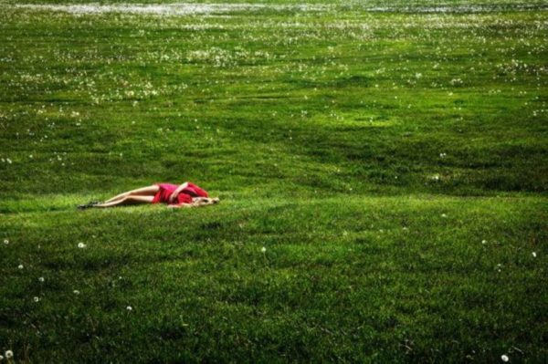 Field of dreams by David Drebin, model in red dress lying in green meadow on the grass