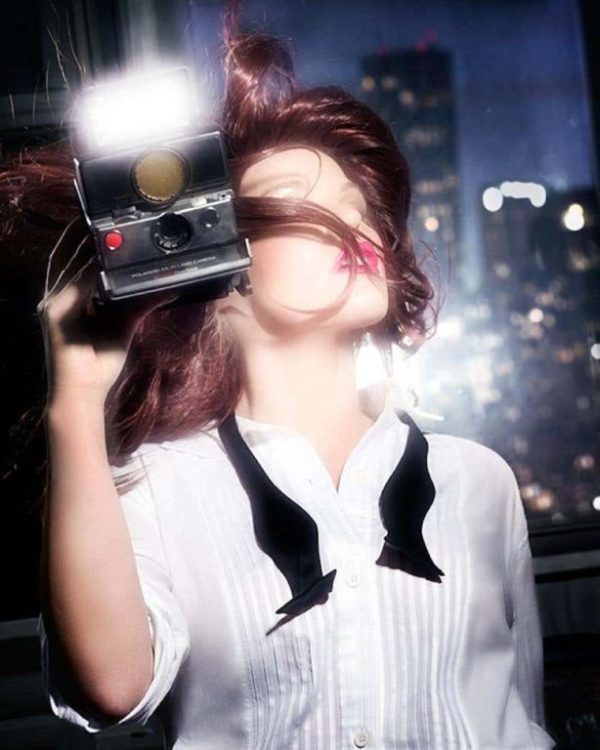 Flashing girl by David Drebin, model in white shirt and open bowtie holding an instant camera and blitzing at the camera