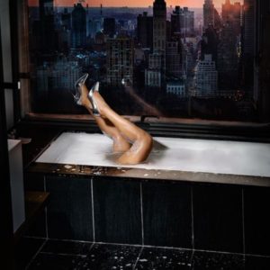 Legging in the city, legs in silver heels sticking out of a bathtub next to a window, outside a city in sunset