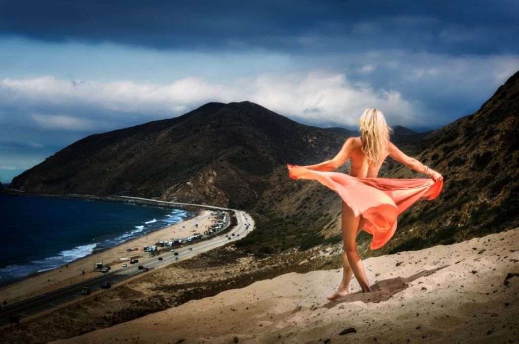 Malibu paradise by david Drebin, model in coral dress twirling on a sandy mountain looking at the ocean