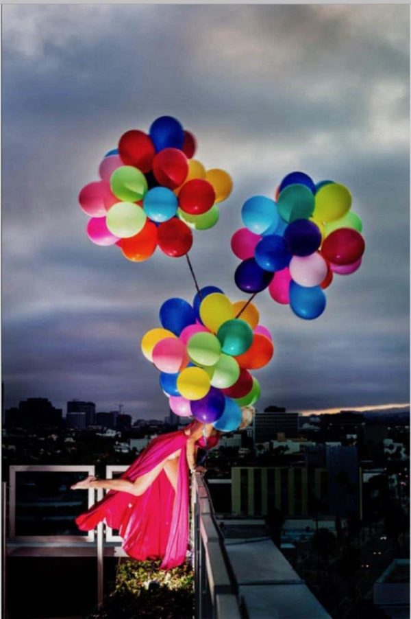 Swept away by David Drebin, model in pink dress, tied to coloful air balloones on a balcony over cityscape