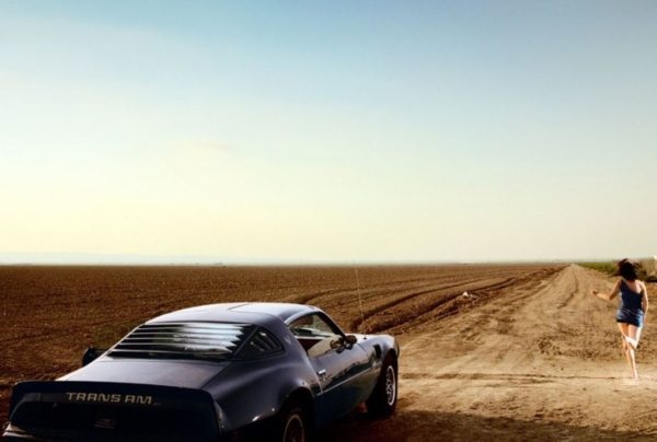 Trans Am by David Drebin, model in short black dress running over an empty field next to a black trans am car