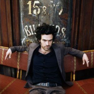 Romain Duris by Gérard Uféras, portrait of the french actor in a brown suit sitting in front of a vintage looking shop