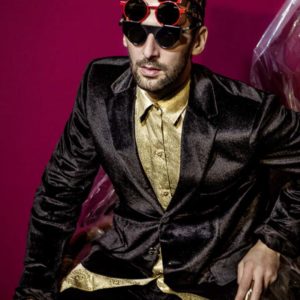 Aaron II by Iris Brosch, male in black suit and gold shirt with several red yellow and black sunglasses on his head