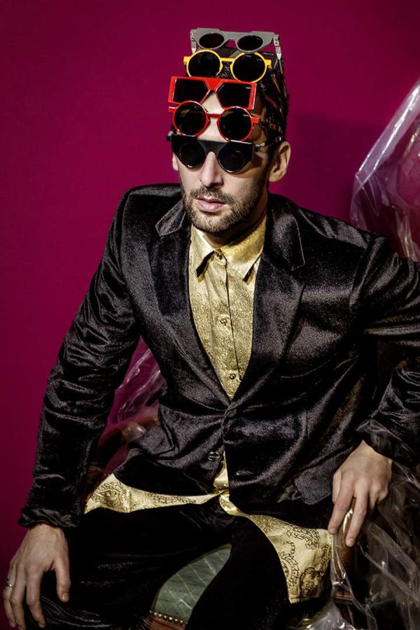Aaron II by Iris Brosch, male in black suit and gold shirt with several red yellow and black sunglasses on his head