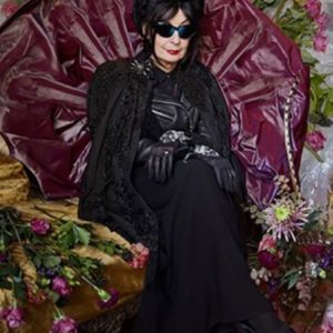 Diane Pernet by Iris Brosch, portrait of the fashion critic wearing all black, sitting inbetween flowers