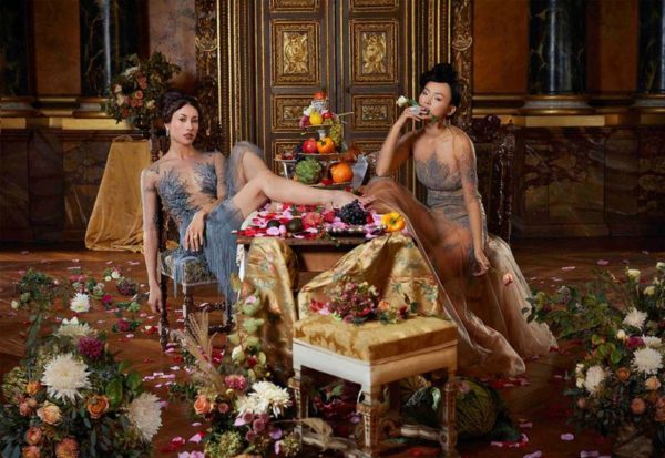 Haute jewelery II by Iris Brosch, two models in transparent gowns lounging at a table full of fruits, surrounded by flowers and petals in a baroque interior