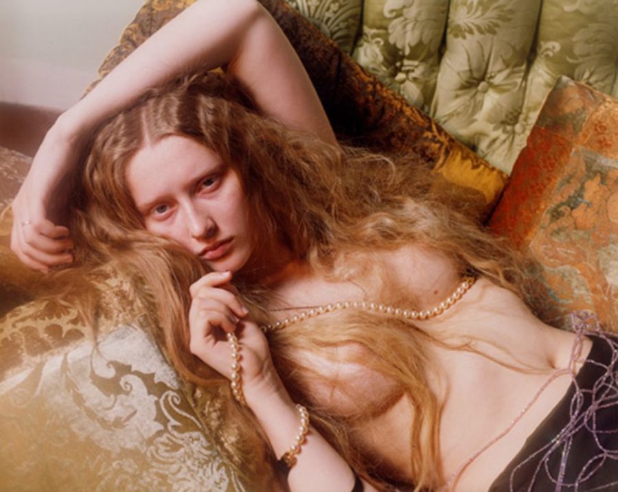 Untitled II by Iris Brosch, portrait of model lounging on a green sofa inbetween orange pillows, her curly blonde hair covering her breasts, a pearl necklase in her hand