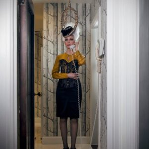 Untitled Magazine by iris brosch, model with white hair updo and fascinator, wearing a black skirt and corset with yellow blouse, standing in a hallway with a cordphone