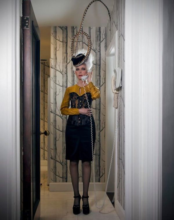 Untitled Magazine by iris brosch, model with white hair updo and fascinator, wearing a black skirt and corset with yellow blouse, standing in a hallway with a cordphone