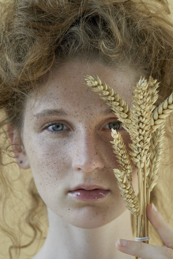 What is Beauty I by Iris Brosch, closeup portrait of model with freckels and messy updo, holding wheat plants in front of her face