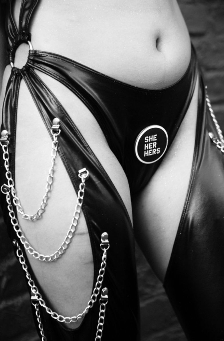 Anaïs Stewart by Marc Baptiste, closeup of a models belly and thighs wearing a leather bodysuit with cutouts and chains and a plaque saying 'She Her Hers'