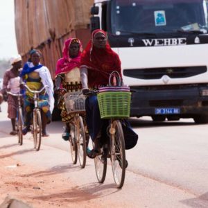 Burkinafaso Ouagadougou II by Marc Baptiste, black women in colorful hidschabs and turbans on bikes next to a truck on the streets