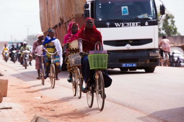 Burkinafaso Ouagadougou II by Marc Baptiste, black women in colorful hidschabs and turbans on bikes next to a truck on the streets