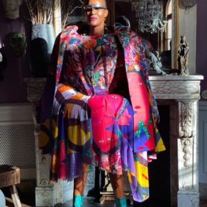 Untitled VIII by Marc Baptiste, model in vuluptous dress and cape with graphic print in pink blue yellow and green, with futuristic glasses and turquoise boots in front of a marble fireplace