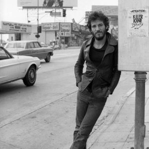 Bruce Springsteen by Terry O'Neill, the musician leaning against a post with cars, a liquor store and a poster in the background