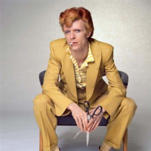 David Bowie 'Yellow Suit' by Terry O'Neill, the singer sitting in a yellow suit with a cigarette in his mout and holding scissors
