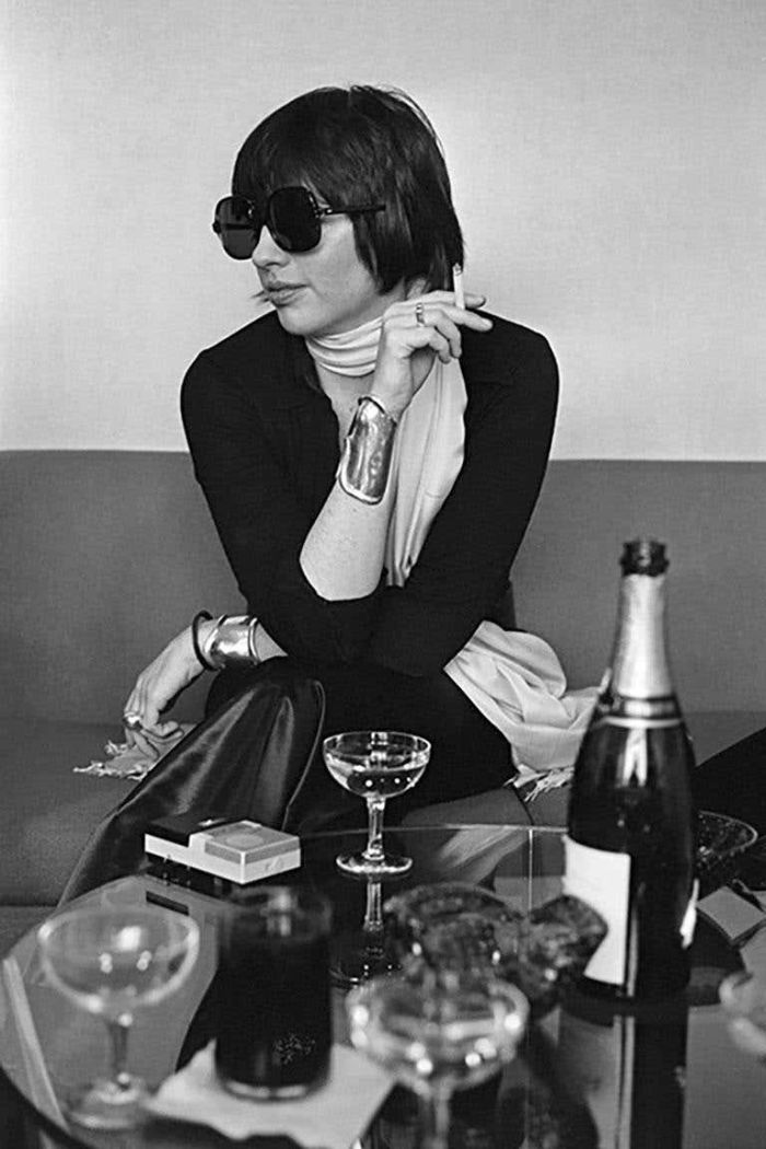 Liza Minnelli by Terry O'Neill, the singer sitting on a couch waerring sungalsses and smoking a cigarette, champagenglasses and bottle in front of her
