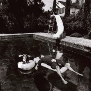 Peter Cook & Dudley Moore by Terry O'Neill, the musicians floating in a pool with floaties, a slide, bushes and a house in the background