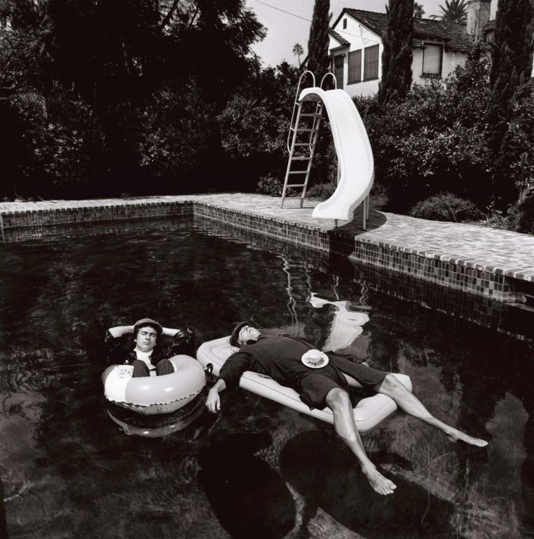 Peter Cook & Dudley Moore by Terry O'Neill, the musicians floating in a pool with floaties, a slide, bushes and a house in the background