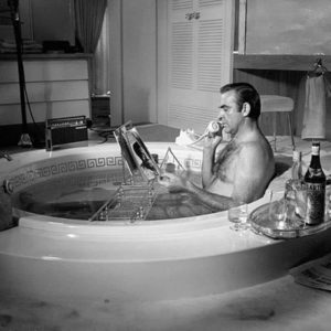 Sean Connery in Bathtub by Terry O'Neill, the actor sitting in a tup next to a Martini, reading a magazine and talking on the phone