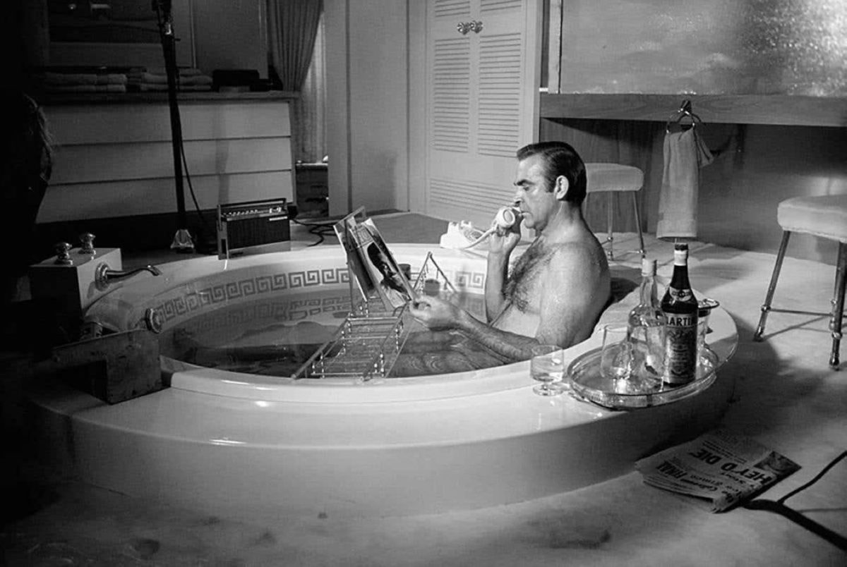 Sean Connery in Bathtub by Terry O'Neill, the actor sitting in a tup next to a Martini, reading a magazine and talking on the phone