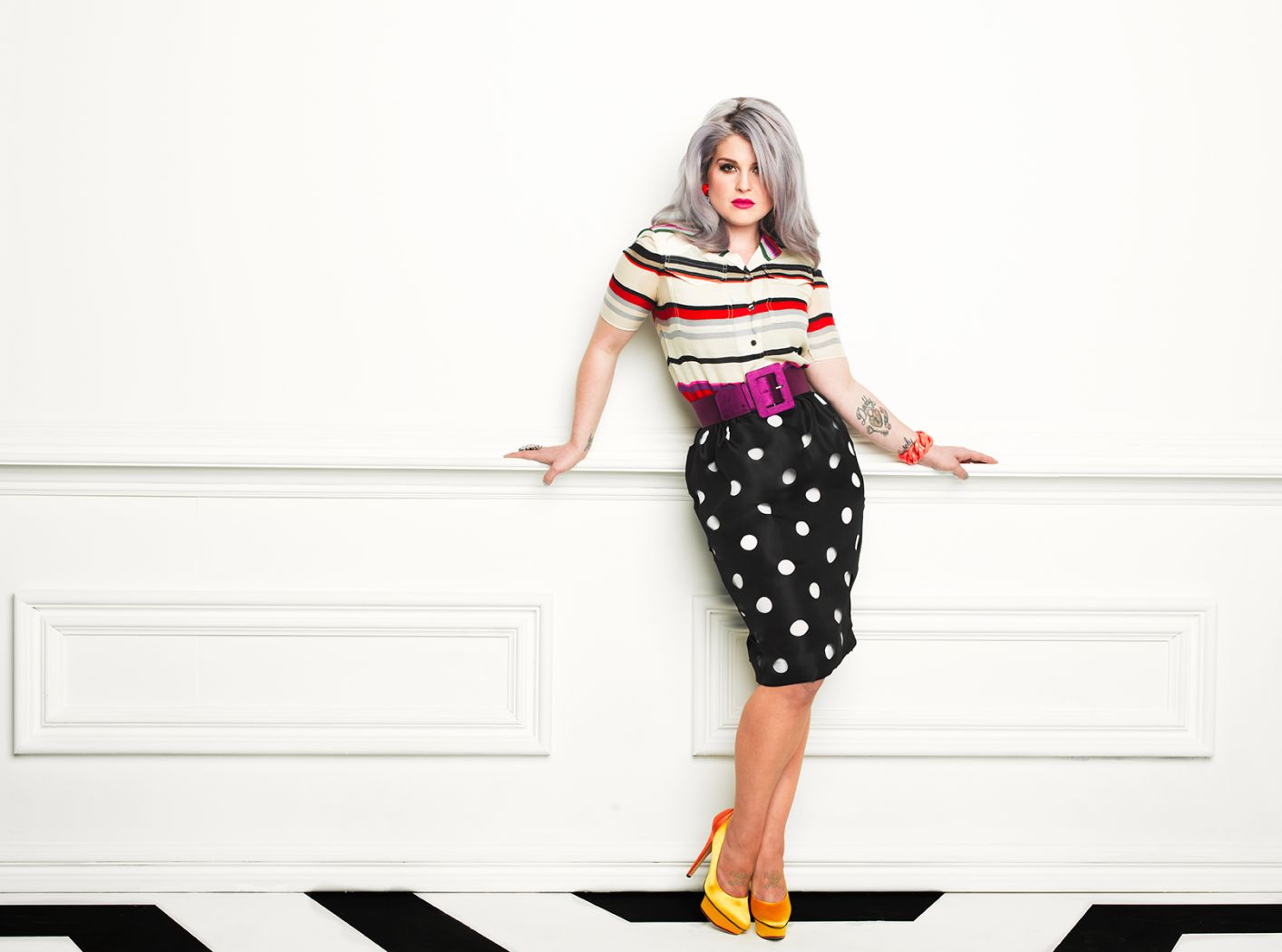 Kelly Osbourne by Timothy white, portrait of the musician in yellow pumps, black polkadot skirt, purple belt and striped shirt, leaning on a wall