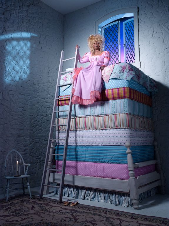 Whoopi Goldberg III by Timothy White, the actress in a pink dress and blonde wig sitting on a stack of colorfull matresses, princess on the pea
