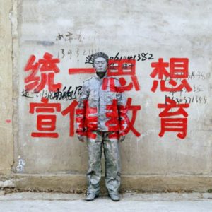 Hiding in the City by Liu Bolin, in front of a graphitty of red chinese signs