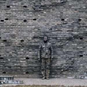 Hiding in the City by Liu Bolin, in front of a grey brick wall