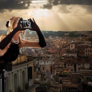 The Great Beauty by David Drebin, model in black with gloves and vintage camera leaning out oif a window over a city scape