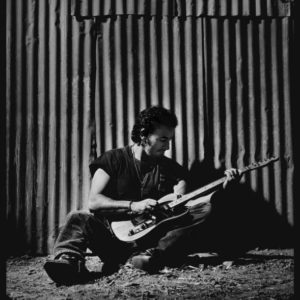 Bruce Springsteen 1992 by Timothy White, the singer sitting in cowboyboots with guitar in front of corrugated iron