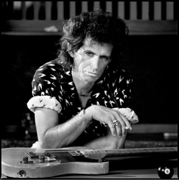 Keith Richards by Timothy White, the musician in a shirt with graphic print, wearing a skull ring and smoking a cigarette, sitting with his guitar