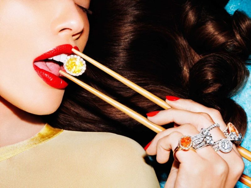 Cosmopola by Tony Kelly, woman with red lip and nails, wearing big rings with orange stones, eating a yellow gemstone with chopsticks