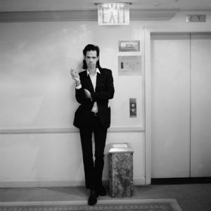 Nick Cave by Jesse Frohman, the musician in a black suit smoking next to a hotel elevator