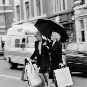 Marilyn and Diana Shopping by Alison Jackson, black-and-white fine art photography of two doubles crossing the street with shopping bags holding an umbrella