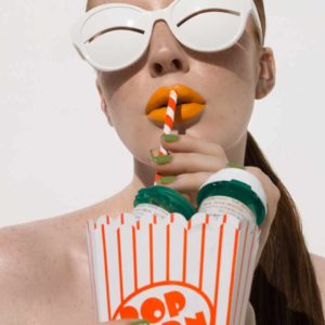"Popcorn, 2022" by Sylvie Blum, from the Space Age Series. Fine Art Print, colour, showing model with brightly coloured lips, wearing white sunglasses, sucking on a straw, holding popcorn bucket
