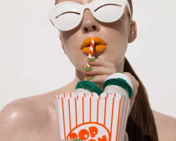 "Popcorn, 2022" by Sylvie Blum, from the Space Age Series. Fine Art Print, colour, showing model with brightly coloured lips, wearing white sunglasses, sucking on a straw, holding popcorn bucket