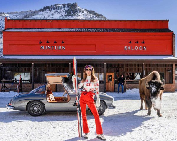 Code Red, Minturn Colorado, 2024 by David Yarrow, Model in red pants with ski standing infront of an oldtimer car, a buffalo and a red Pub