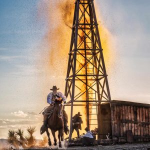 The Golden age of Oil, 2023 by David Yarrow - an old cowboy galopping towards the Camer, in the background an exploding oil rig