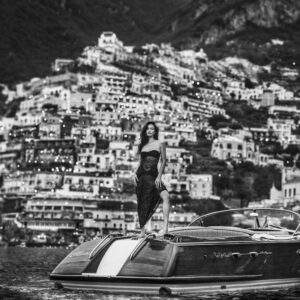 La Dolce Vita Positano by David Yarrow - Model standing on a Motorboat infront of the italian Coastline with coastal village in the background