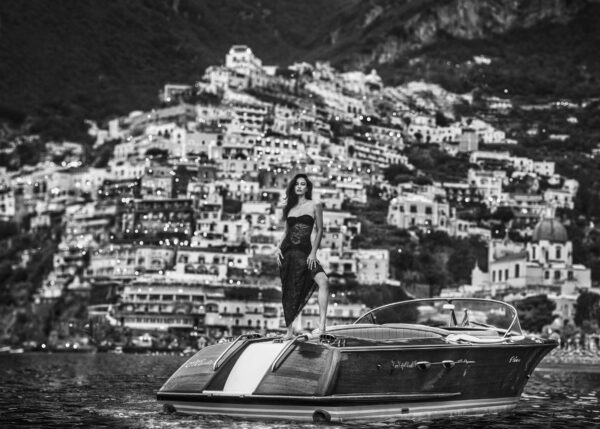 La Dolce Vita Positano by David Yarrow - Model standing on a Motorboat infront of the italian Coastline with coastal village in the background