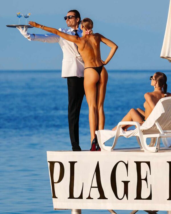 La Dolce Vita I, Plage Keller, Cap d’Antibes 2024 by Tony Kelly, nude model reaching for a drink a waiter in a suit is holding next to the ocean on the sun deck of the Palge Keller