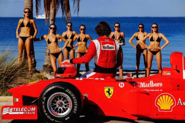 Pit Stop 2024 by Tony Kelly, Formula one driver on his red Ferrari Race Car looking at Models standing by the beach.