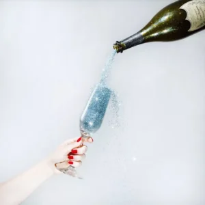 Tyler Shields Glitter Champagne, glitter pouring from a champagne bottle into a glass held by a womans hand with red nailpolish